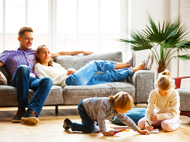Family in a sunny living room, parents relax on a sofa while two young children play on the floor.