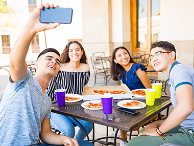 Group of four young people sit around a metal outdoor patio table with pizza and drinks in plastic cups; they're taking a selfie.