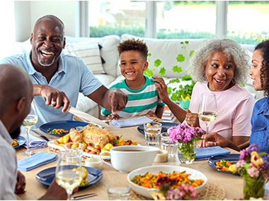 Three generations of a Black family, talking happily around an intimate dining table laden with a feast, in a light-filled dining room.