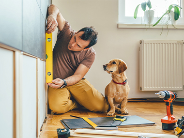 Man works on a home improvement project, checking a wall with a level, while his dog looks on.
