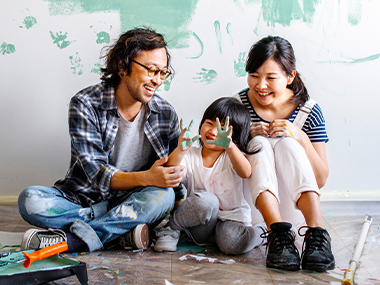 Casually-dressed smiling Asian family, sits on the floor, taking a break from painting a wall; young girl holds up her hands, covered with teal paint.