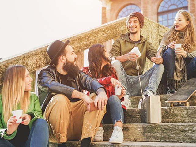 A mixed group of college students sit on campus steps while talking and drinking beverages
