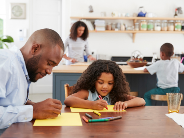 An African-American father and daughter draw on kitchen table with mother and son in the background