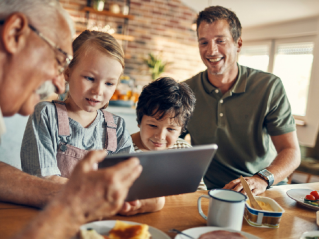 A grandfather holds a tablet as his son and 2 grandchildren view the screen