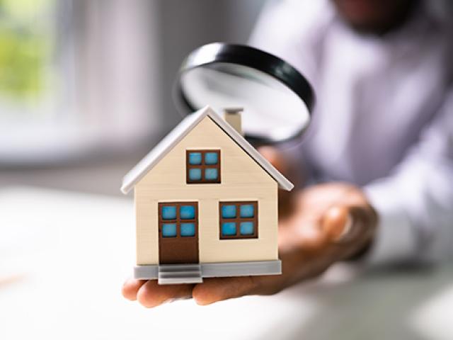 A man's hand holding a small model of a home while he inspects it with a magnifying glass