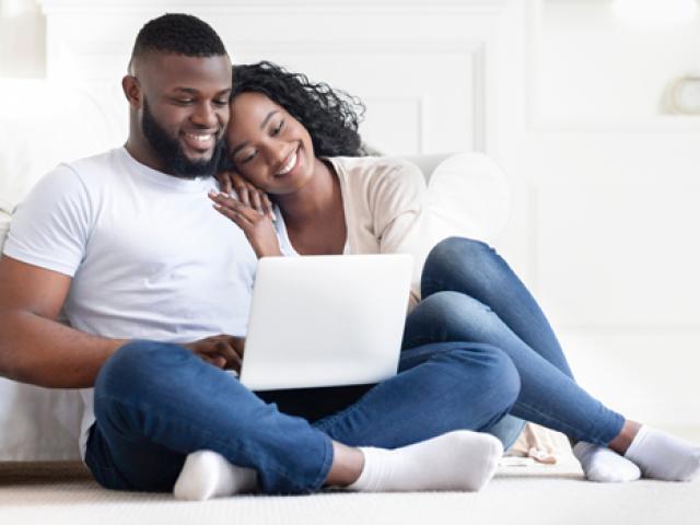 A couple sitting on the floor while using a laptop and smiling