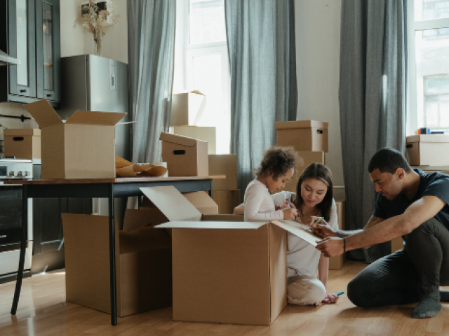 A couple sit on the floor with their preschool daughter surrounded by moving boxes 
