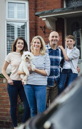 Smiling family of four with two pre-teens and a white poodle, stand in front of a brick home.